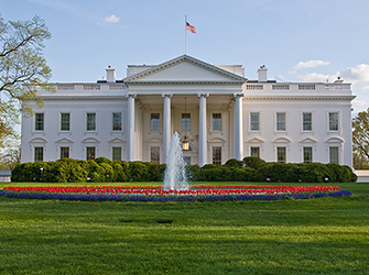 There is a two story white building with columns. There is an American flag on top of the building. In front of the building is a large yard with a fountain surrounded by red and blue flowers.