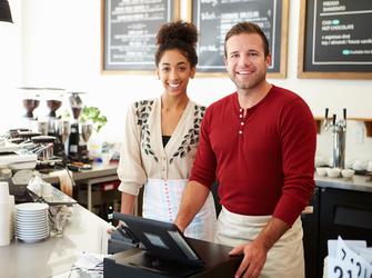 A woman and man wearing aprons stand smiling behind the serving counter at a coffee shop.